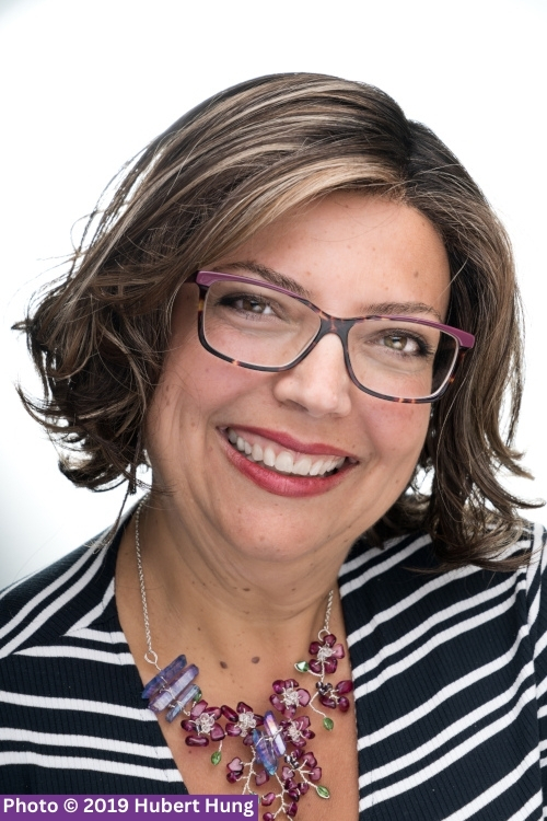 Frederika Roberts - middle-aged woman with a big smile, dark slightly wavy hair in a bob, with highlights, and burgundy and tortoiseshell glasses. Frederika is wearing a black and white striped top and a chunky colourful necklace with lots of pink.